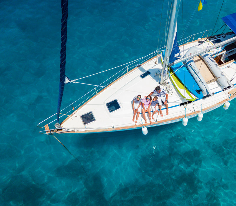 A family sitting on a yacht over clear blue water, looking up at a drone taking an aerial photo